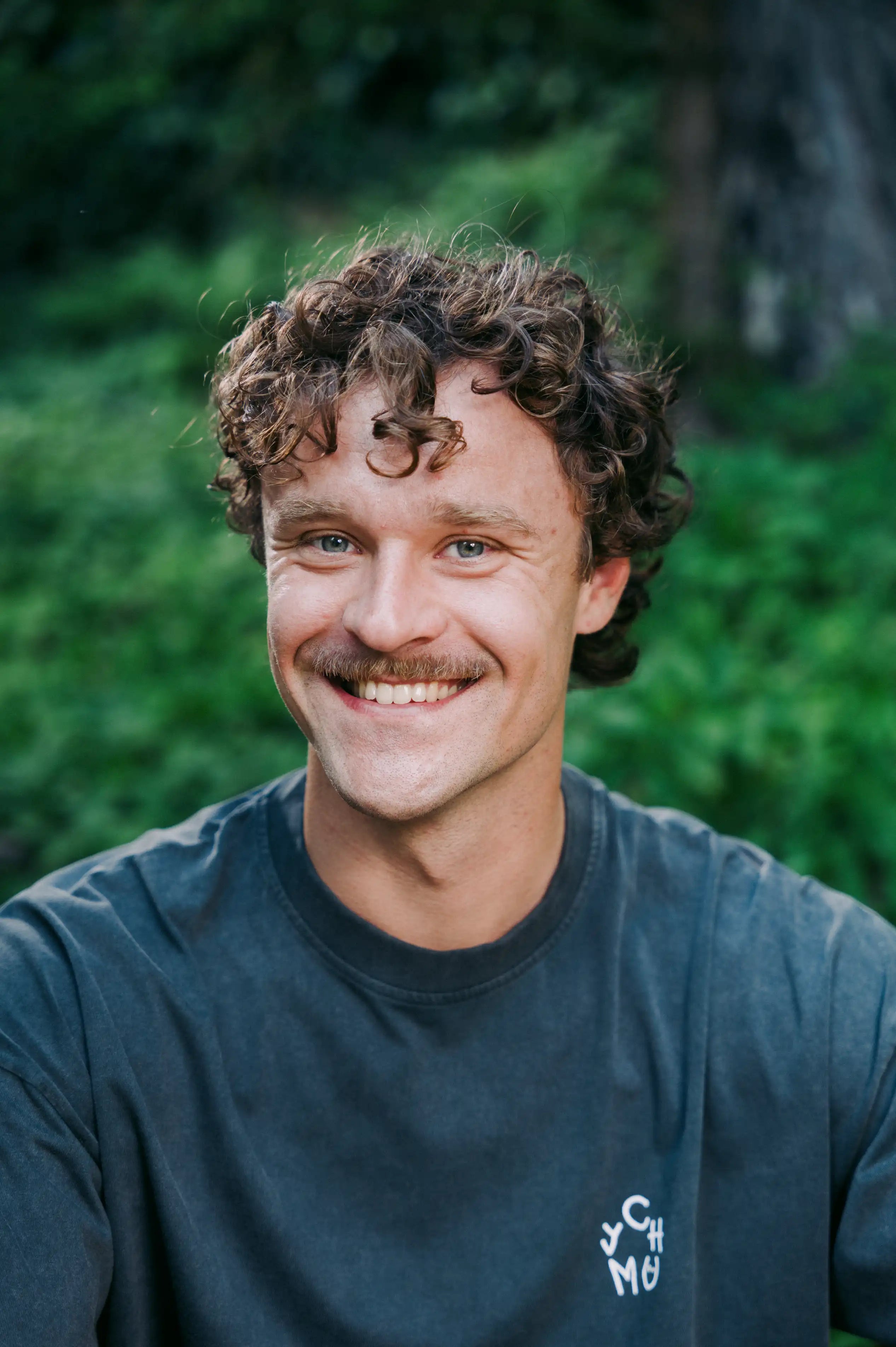 Nahaufnahme eines lächelnden Mannes mit lockigem Haar und Schnurrbart im Chumy-Shirt. Das Team hinter der Sportnahrung für Laufen, Triathlon und Radsport.