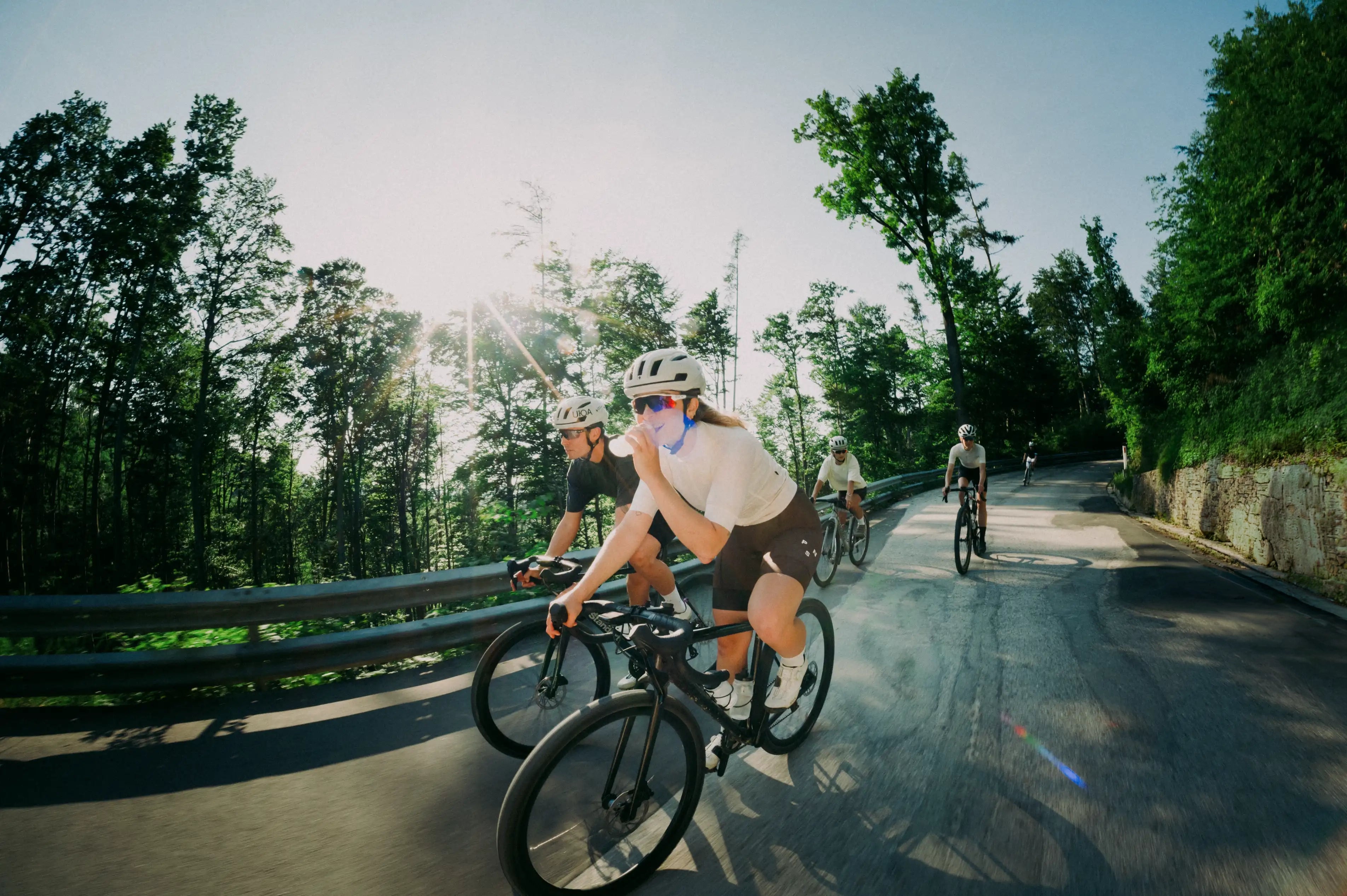 Eine Radfahrerin trinkt im Fahren auf ihrem Rennrad bei Sonnenschein. Dahinter folgt eine Gruppe beim Ausdauersport auf einer kurvigen Straße am Waldrand. 