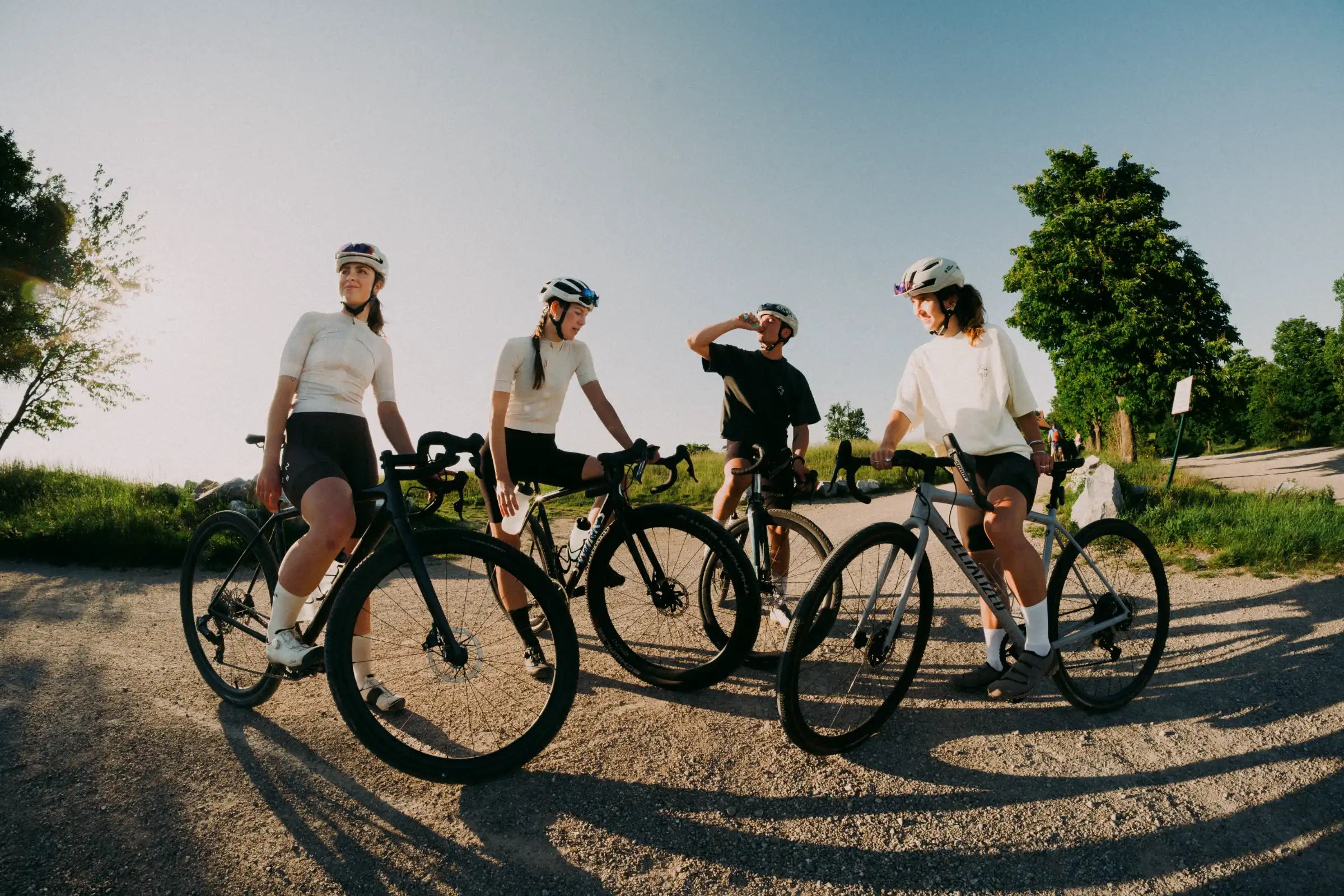 3 Radfahrerinnen und ein Radfahrer stehen an einer Lichtung auf einer Schotterstraße im Sonnenuntergang. Sie konsumieren nachhaltige Energie Gels und Elektrolyte